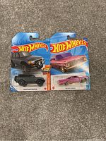 Photo shows two Hot Wheels die-cast cars factory sealed in original blister packaging on blue Hot Wheels cards. Left car is gray Toyota Land Cruiser FJ, right car is purple Chevy Impala with flame decals.