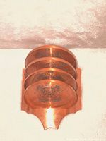 Set of four round copper coasters with engraved emblem stacked inside a matching copper holder with scalloped front edge on a light fabric surface.