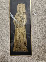 Full view of the framed brass rubbing artwork showing a medieval figure in robes with hands in prayer position, mounted on a dark background with golden frame.