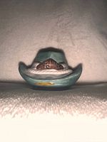Photo of cowboy hat shaped lint remover brush on a white textured surface, showing front angle with leather strap detail.
