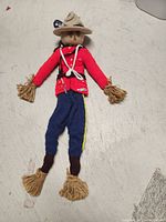 Full-length photo of the knitted RCMP Mountie doll lying flat, showing red tunic, blue pants with yellow stripe, brown boots with fringed yarn ends.