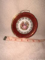 Front view of the small porcelain plate with decorative border and central scene, showing approximate size with measuring tape.