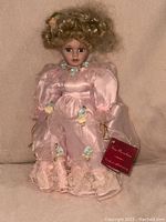 Porcelain doll 14 inches tall, in pink satin dress with lace and floral decoration, attached stand visible, curly blonde hair, attached tag.