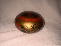 Side view of the round wooden trinket box featuring gold paint with red and black floral folk art patterns.