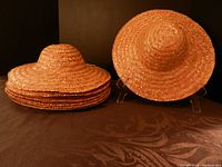 Ten tan mini straw hats stacked and displayed on stands on a black surface and background, clearly showing the shape and size of the hats.