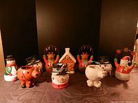 Group photo showing ten assorted ceramic Christmas character votive holders and four glass snowman votive holders arranged on a surface with a dark background.