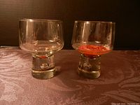 Two clear glass candle holders with thick stems and round bowls on top, one containing orange wax residue