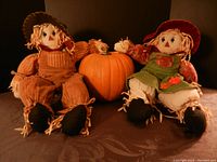 Both stuffed scarecrow dolls seated with faux pumpkin between them on a dark surface with black background