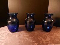Three cobalt blue glass vases shown side by side on a textured fabric surface, front view highlighting bulbous body and narrow neck with flared rim.