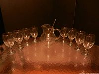 Glass beverage set including one pitcher with handle and acrylic stirrer, with eight clear glass goblet-style beverage glasses arranged around it on a reflective surface.