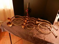 Overview of nine brass horns of varied sizes including large and small round horns and the straight horn with red string tied on neck, displayed on a table with dark patterned cloth.
