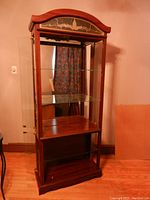 Full front view of the tall wood display cabinet featuring glass shelves and mirrored back inside, and decorative arched top panel.