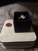 Diamond aura infinity ring in silvertone inside black ring box placed on a beige jewelry box, penny nearby for size comparison.