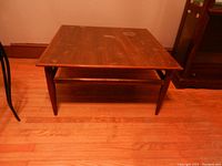 Square brown wooden mid-century modern coffee table viewed from side and slightly above. Shows tapered legs and lower shelf.