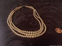Full view of a 3-strand faux pearl necklace laid flat on a black textured surface next to a penny for scale