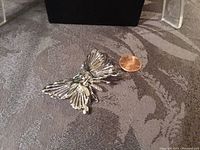 Silvertone en tremblant butterfly brooch angled showing detailed wings and green crystal eyes with penny for size reference.