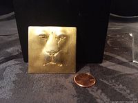 Front view of the square goldtone metal lion face brooch next to a penny for size reference.