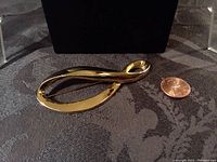 Front view of the goldtone loop brooch placed next to a penny for size reference, showing smooth shiny finish and loop shape.