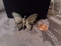 Front view of the gray metal butterfly brooch placed beside a penny for size reference, showing the detailed wings and body.