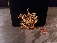 Front view of goldtone lily of the valley floral brooch showing detailed flowers and leaves next to a penny for size.
