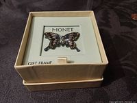Butterfly pin centered in Monet gift frame box, showing front details and box label, on dark fabric background.