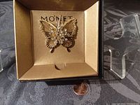 Front angled view of vintage Monet butterfly pin inside box with penny for scale, showing goldtone finish and crystal accents.
