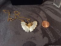 Photo of butterfly pendant necklace with mother of pearl wings next to a penny for scale, showing the front side.