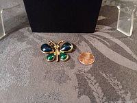 Front view of a goldtone butterfly brooch with blue and green painted wings and white crystal eyes, placed beside a penny for scale.