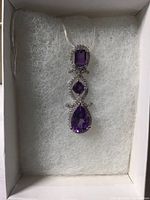 Pendant displayed in white cushioned box showing rectangular, square, and pear cut amethysts surrounded by white cubic zirconia.