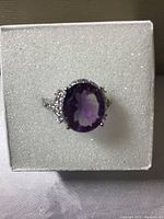 Top view of oval cut deep purple amethyst ring with white cubic zirconia stones around it, set in 14k white gold over 925 silver, placed inside a cushioned box.