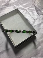 Photo showing entire bangle inside white cushioned box highlighting the green oval diopside stones and metallic band.