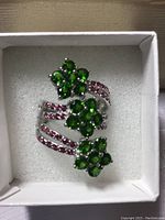 Ring displayed upright in white jewelry box showing three green flower clusters and ruby-set band.