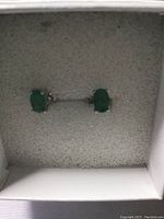 Pair of oval emerald stud earrings set in 925 sterling silver displayed in white jewelry box with foam insert.
