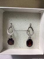 Front view of pair of oval garnet drop earrings with white CZ accents in white gold over silver, displayed in a white box.