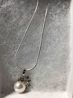 Close-up photo of 10mm white pearl pendant with multiple small colored gemstones including citrine, diopside, and red garnet on 14k white gold over 925 setting, hanging from fine chain inside a box.