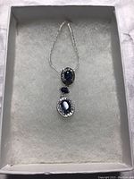 Pendant with two oval blue sapphires and white CZ stones on a silver chain inside a white box.