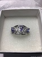 Ring with three pear-shaped blue-violet Tanzanite stones set in 14k white gold over 925 sterling silver, accented with white cubic zirconia stones displayed in white jewelry box.