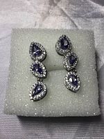 Photo showing both earrings laying flat on white foam, displaying the shape and arrangement of the pear cut tanzanite stones and CZ halo.