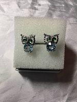 Pair of owl earrings displayed on white foam with visible blue topaz gemstones and green eyes