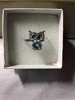 Owl ring featuring blue topaz gemstone body, green diopside eyes, white cubic zirconia accents, in white gold plated sterling silver mount, shown in jewelry box.