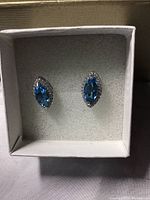Top view showing pair of blue marquis cut topaz stud earrings surrounded by white cubic zirconia in a white box.
