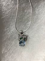 Close-up photo of owl pendant hanging from chain on grey foam background. Shows blue Topaz on owl body, green Diopside eyes, and white CZ accents in white gold over silver setting.