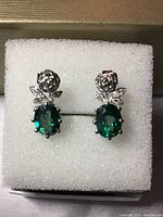 Close-up image of pair of earrings with green oval cut stones and white CZ shaped like roses and leaves, set in white gold over silver, displayed on white foam in box.
