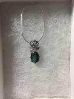 Front view of a rose pendant with an oval cut green topaz and white cubic zirconia accents on a chain, displayed on white padding inside a box.