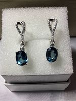 Photo showing pair of dangle earrings with oval London blue topaz gemstones and white cubic zirconia accents on white foam padding inside box.