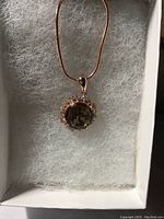 Pendant resting in a white cushioned box, showing round smoky topaz center with surrounding white cubic zirconia and rose gold chain.