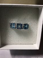 Close-up photo of the ring showing three square princess cut Swiss Topaz stones in a row, light blue in color, mounted on a white gold over silver band, set in a white foam cushion in a box.