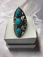 Front view of a silver ring with three turquoise stones set vertically in decorative filigree silverwork on a white foam cushion inside a white box.