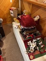 Stacked Christmas decorations including teddy bear in red clothing, Santa Claus doll, trays of ornaments, fabric poinsettia flowers, and holiday table runners.