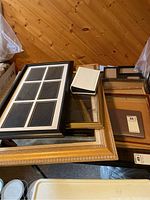 Table with a stack of assorted picture frames in various sizes and styles, showing different frame colors including gold, black, and natural wood. One large frame at the bottom with missing glass.